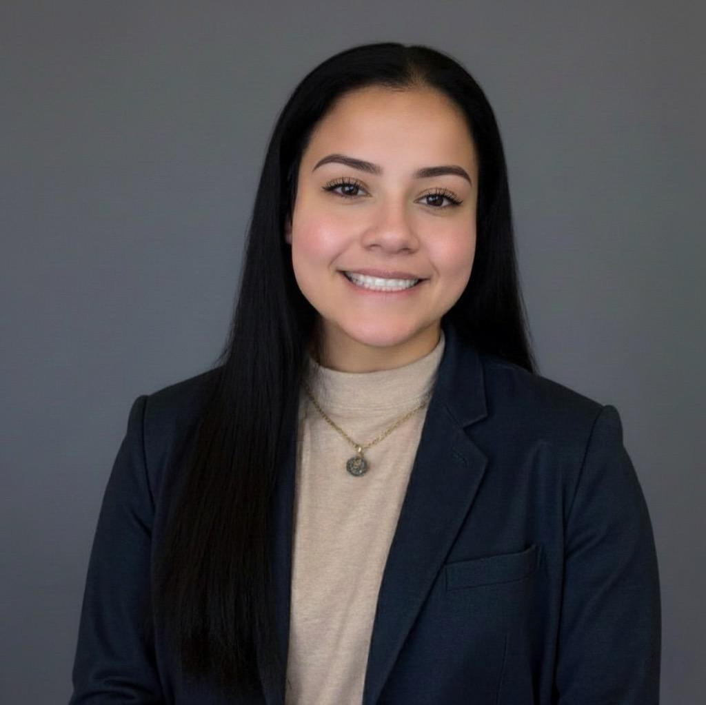 The image is a headshot of Gabriela Martinez, co-founder and CEO of Tekcesta LLC. She is wearing a dark blue blazer over a tan shirt, casually dressed. Gabriela is a 23-year old Hispanic American woman. She has long brown hair and brown eyes. She is smilin